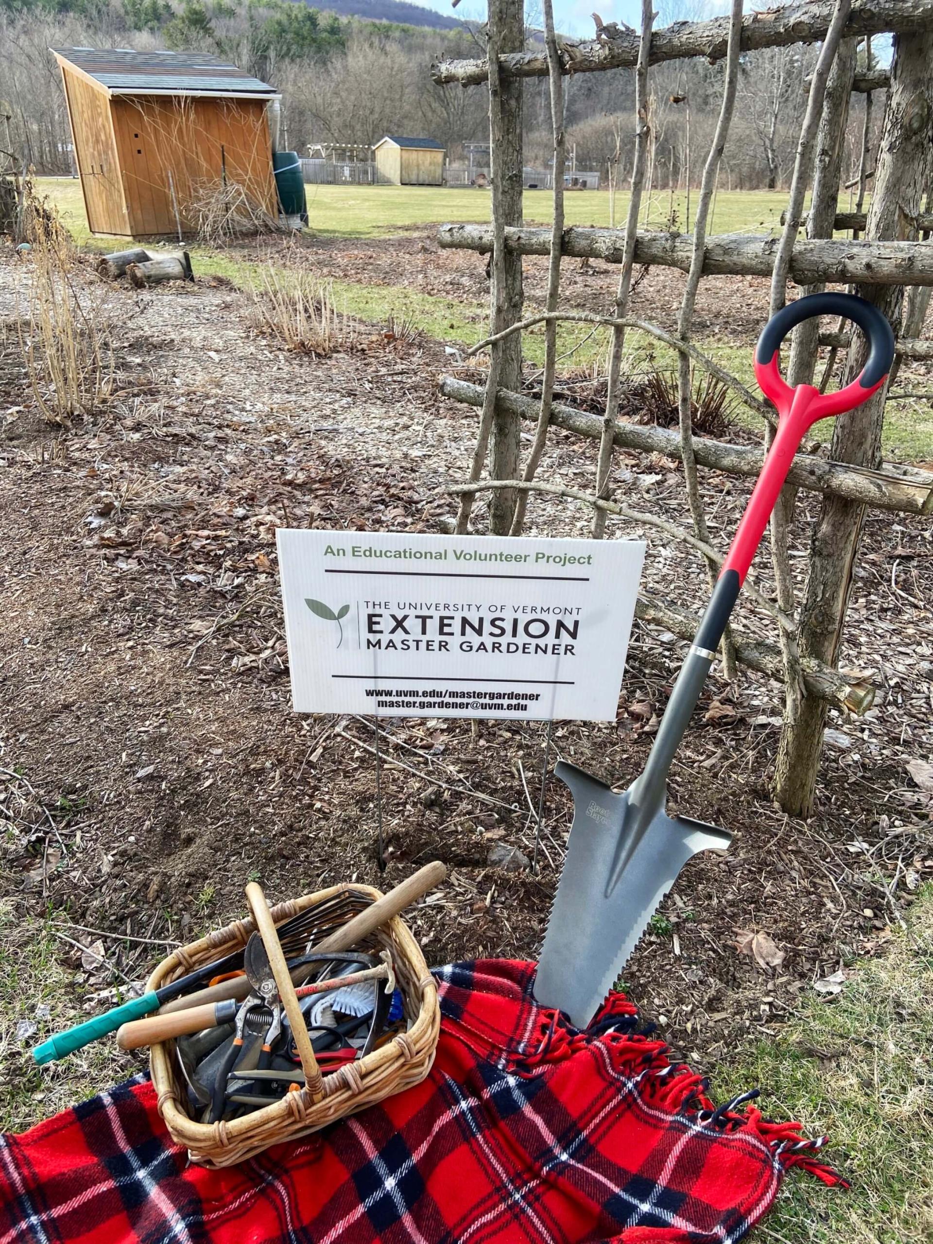 A shovel next to a basket full of garden tools on a plaid, red blanket
