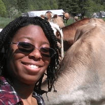 image description: a smiling young woman with sunglasses leaning against the neck of a brown cow on a sunny day