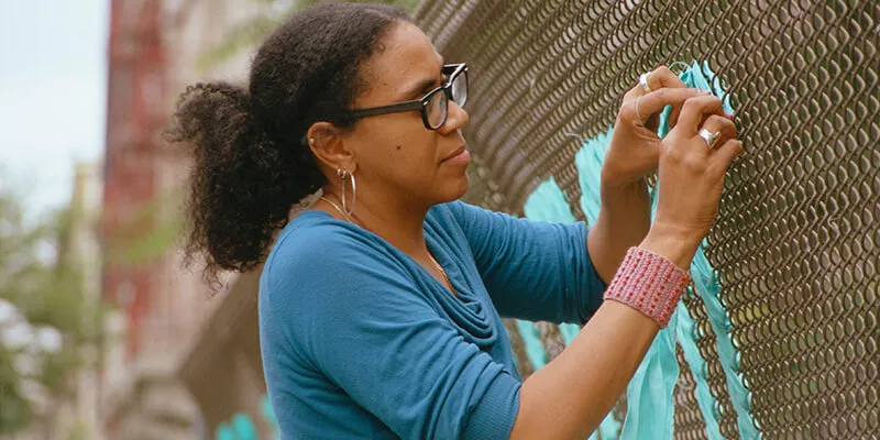 Mildred Beltre weaving material into a Brooklyn roadside metal fence.