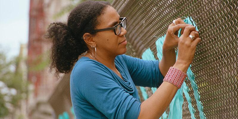 Mildred Beltre weaving material into a Brooklyn roadside metal fence.