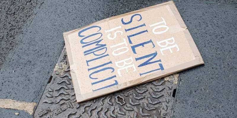 protest sign on street reads "To be silent is to be complicit"