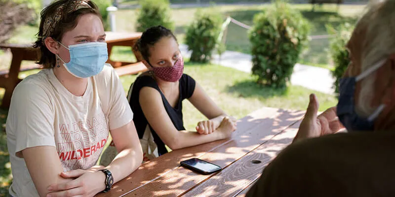 Two students interview a man at a picnic table.