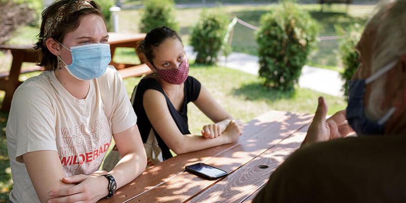 Two students interview a man at a picnic table.