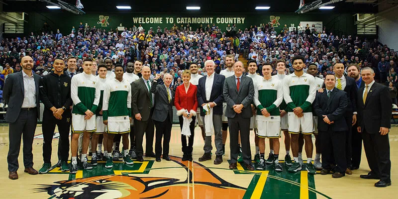 Men's basketball players and university leaders join Rich and Deb Tarrant on the basketball court with fans in bleachers behind them