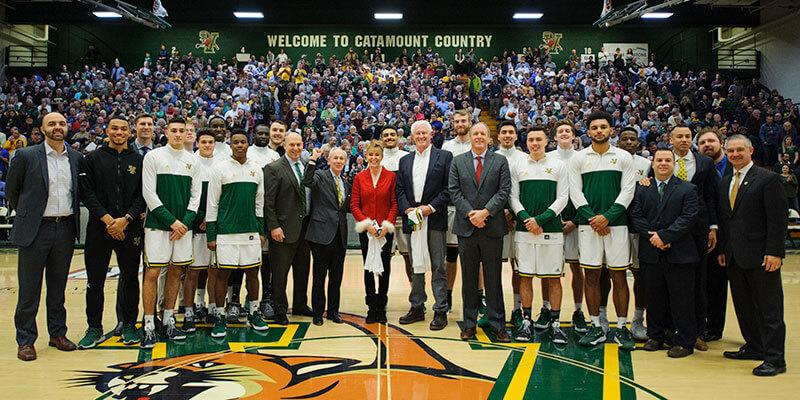 Men's basketball players and university leaders join Rich and Deb Tarrant on the basketball court with fans in bleachers behind them