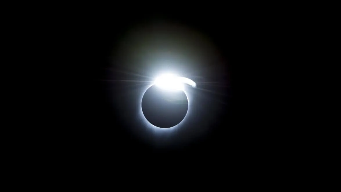 An image of a solar eclipse, with the sun almost entirely blocked by the moon, with a small amount of light coming through. The background is black.