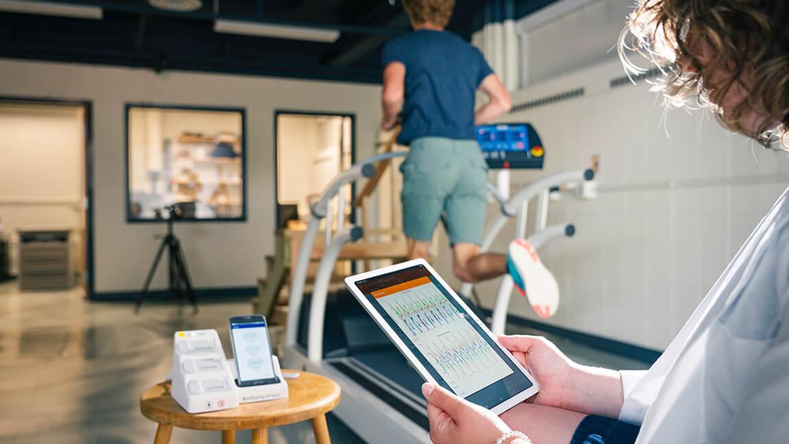 A practitioner looks at data on a tablet while a patient jogs on a treadmill