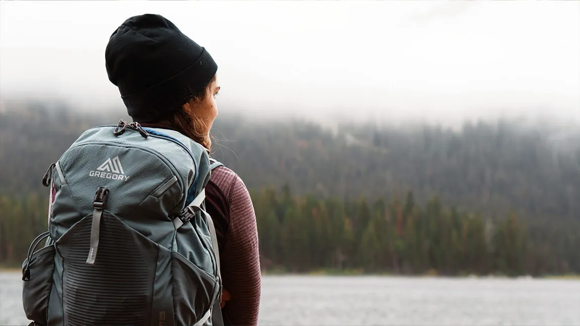 teenager wearing a hat and a backpack staring out over a lake towards some dense woods