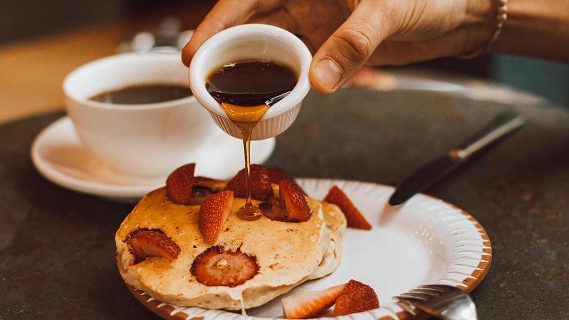 someone pouring maple syrup from a small ceramic container onto pancakes