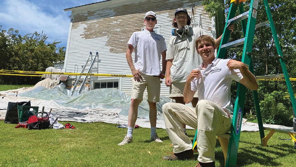 three students in white polos stand in front of a partially painted house, caution tape, a ladder, and other painting equipment