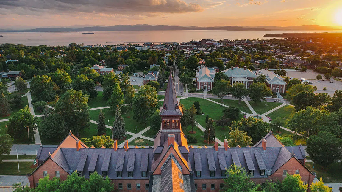 An aerial photo of the University of Vermont's historic campus overlooking the city of Burlington and Lake Champlain with the sun shining. 
