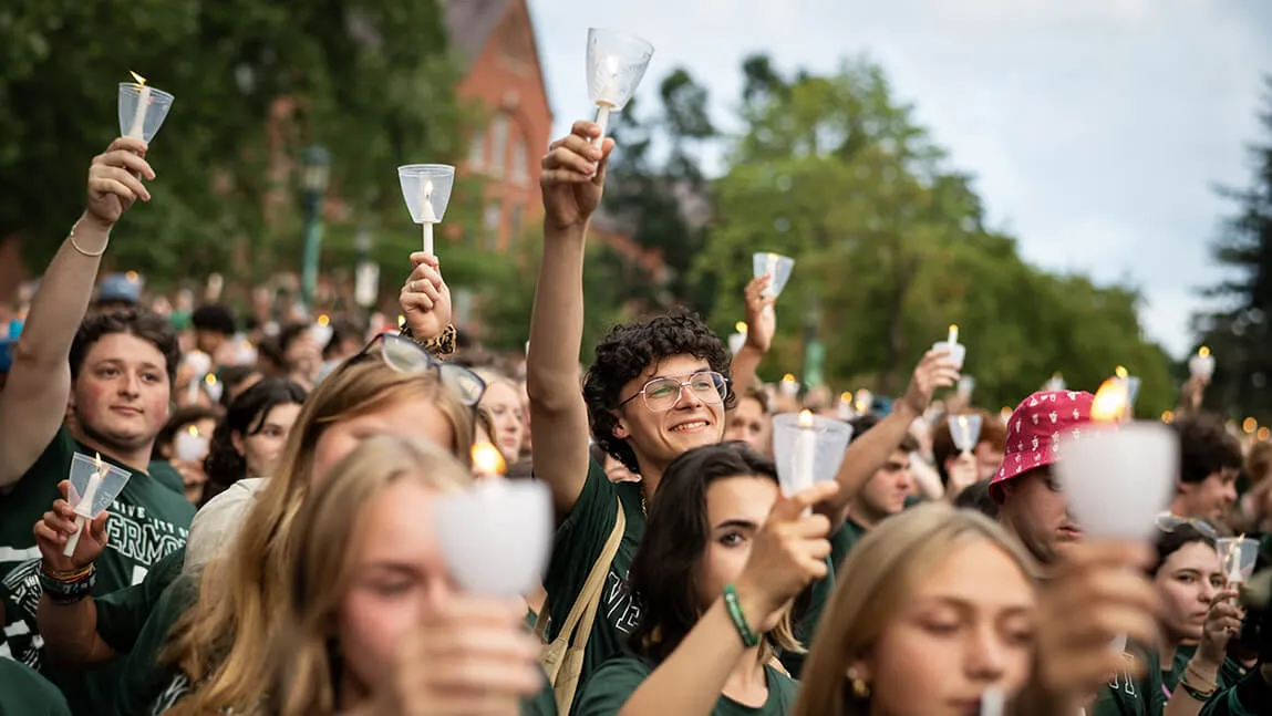 students in a crowd smile while holding lit candles in the air in front of the backdrop of old mill on the university green