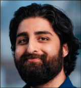 A man with dark brown hair and a beard wearing a blue shirt