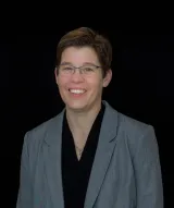 Person with short hair wearing glasses and a black top and gray blazer, facing the camera against a black background