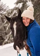 Brown haired woman in blue sweater with a cream colored hat standing outdoors in the snow with a black horse