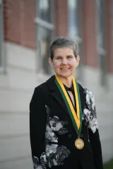 Person wearing a black floral-patterned jacket and a gold medal on a green-and-gold ribbon, standing outdoors in front of a blurred building.