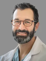 Professional headshot of a person wearing rectangular eyeglasses and a light-colored collared jacket over a blue shirt. The person has short, dark, wavy hair and a full beard and mustache. The image is cropped from the shoulders up and set against a plain gray background, with the subject facing the camera in a formal portrait style.