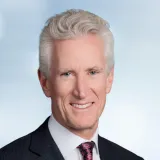 Professional headshot of a person with short gray hair wearing a dark suit jacket, white dress shirt, and patterned red tie. The person is facing the camera against a light blue gradient background. The image is cropped from the shoulders up and has a formal business portrait style.