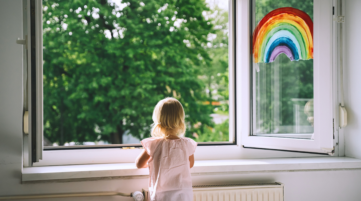 Little girl looks out open window at spring time. Painting rainbow on window. Open window in room with green trees on background. Kids leisure at home. Support during quarantine. Feeling fresh air.