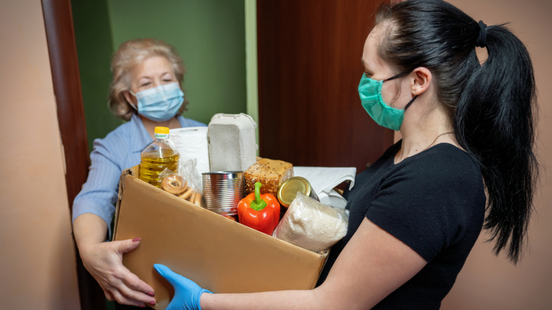 Volunteer young female in medical mask and gloves handing an senior woman a box with food. Donation, support people in quarantine, coronavirus