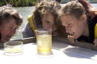A teacher and two students in yellow life vests examine a water sample in a beaker.