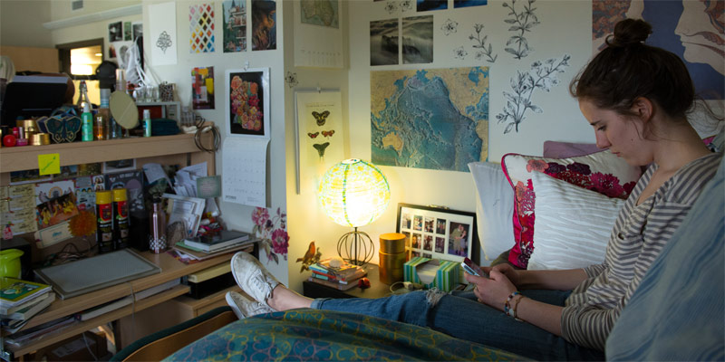 A student sits on the bed in her dorm room and checks her phone