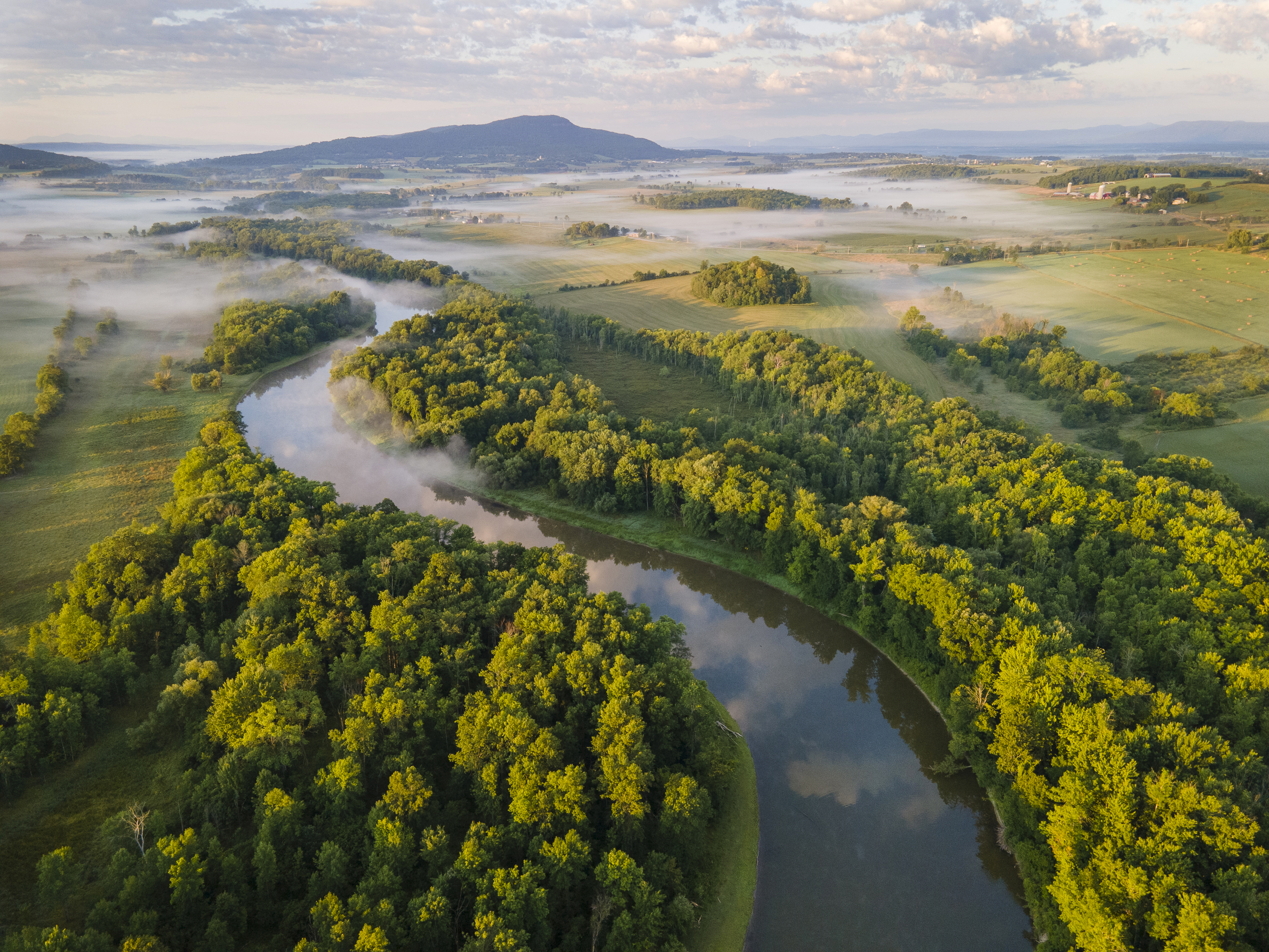 View of Camel's Hump taken from drone, with farm fields and forest buffers around Winooski River on a day with morning fog.