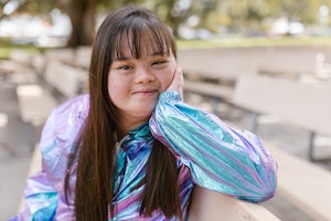 A teenage Asian girl with Down Syndrome sits on a bench in an open park. She rests her chin on one hand and smiles openly at the camera.