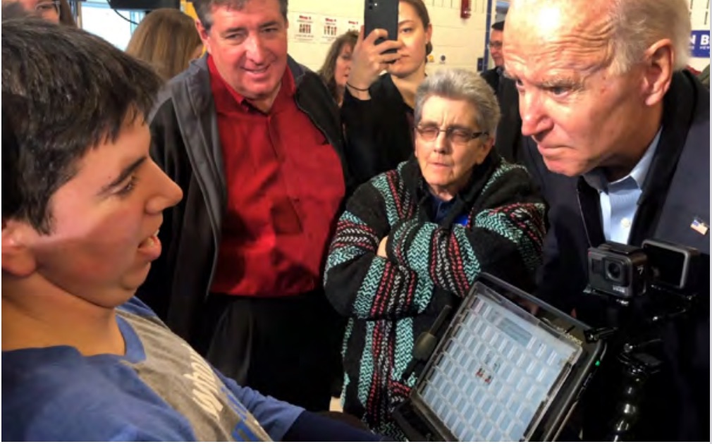A teenager in a wheelchair, with an adaptive communication device in front of him, meets the gaze of President Joe Biden, as onlookers watch and film them with their cameras.
