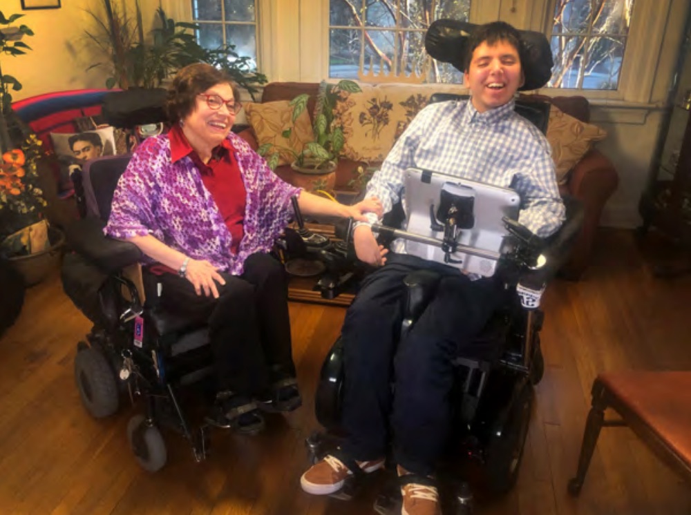 A teenager in a wheelchair sits in a warm front parlor next to an older woman in a wheelchair. He is smiling broadly, and she wears a bright purple shawl.