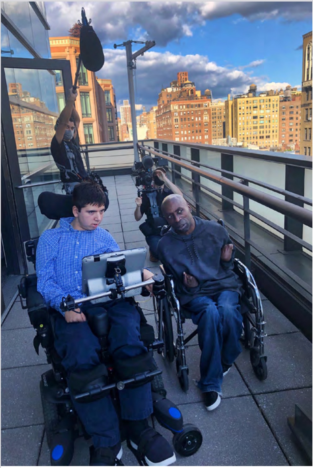 A teenager in a wheelchair sits on the balcony of a skyscraper with a young Black man in a wheelchair. Behind them, a masked man holds a boom mic over their heads.