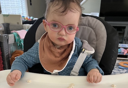 A toddler in shell-pink glasses sits in a high chair and regards the camera dubiously, unsmiling.