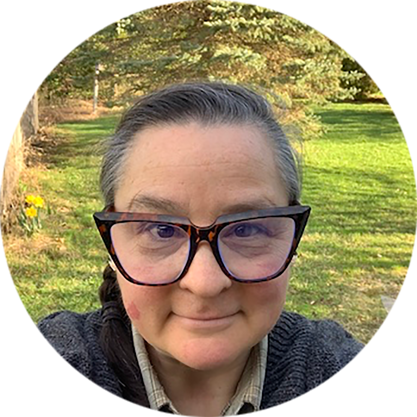 Audrey Homan: a white non-binary person in their forties, dark hair pulled into a braid over one shoulder, and wearing large, heavy-framed glasses, staring directly into the camera in front of a sunny evergreen.