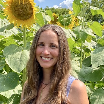 Amy Starble: a pale-skinned woman in her mid forties, with long brown wavy hair worn loose and down, smiles invitingly in front of a sunlit field of sunflowers. A large, open sunflower hangs over her head.