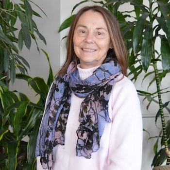 Phoebe Chestna: a white woman in her late sixties, with straight gold-brown hair worn loose, stands in front of greenery in an atrium. She wears a purple floral scarf draped loosely around her neck.