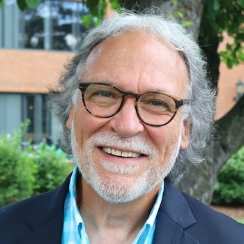 Michael Giangreco profile photo: an older white man with short white hair, glasses, and full white beard and mustache stands smiling proudly in front of a red brick building and a lush green tree.
