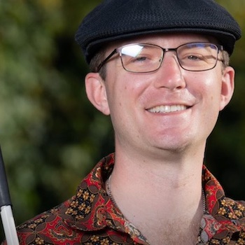 Justin MH Salisbury: a mixed race man with pale features, in his early thirties, wearing wire-framed glasses and a flat cap, smiles confidently at the camera, holding a white cane against his shoulder.