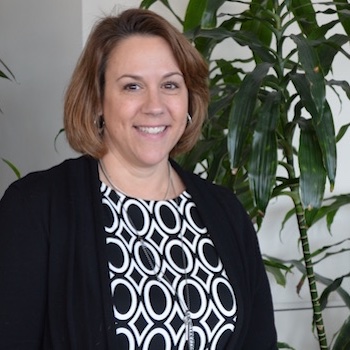 A smiling white woman with wavy, honey-brown hair in a bob, stands in front of a ficus tree and fireplace. She is wearing a smart blazer and checkered work blouse. Valerie LeClair