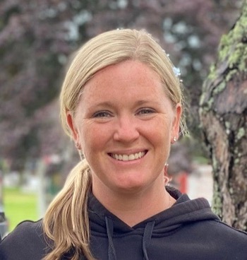 Nicole Howrigan, a blonde white woman in her late twenties, smiles confidently at the camera. Her hair is pulled back in a loose ponytail and she stands outside, in front of a tree trunk.