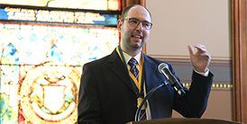 Professor Matthew Price with investiture medal  at a podium with stained glass window in backgroun