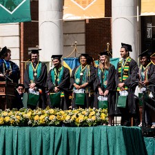 Six students in academic regalia stand in a row across the commencement stage. In the foreground is a row of green and yellow flower arrangements lining the front of the stage, which is has green brocade pleated fabric skirting. In the background hang large banners between giant columns of the brick building behind.