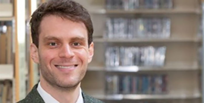 Giacomo DiPasquale smiling broadly, indoors with booklined bookshelves in the background