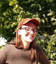 image description: woman with brown hair and glasses and an orange baseball hat, smiling towards the camera, in bright light and in front of leafy green trees