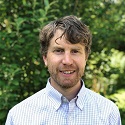 Headshot of Gregory Rowangould. He is smiling in front of greenery outdoors