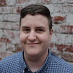Robyn in front of a faded brick wall wearing a blue checked shirt and smiling