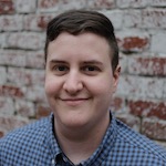 Robyn in front of a faded brick wall wearing a blue checked shirt and smiling