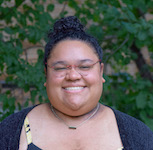 Kay in front of a dark green leafy background, smiling towards the camera, with a black sweater and sundress on