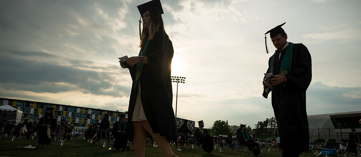 Two UVM graduates silhouetted against sun walk in caps and gowns