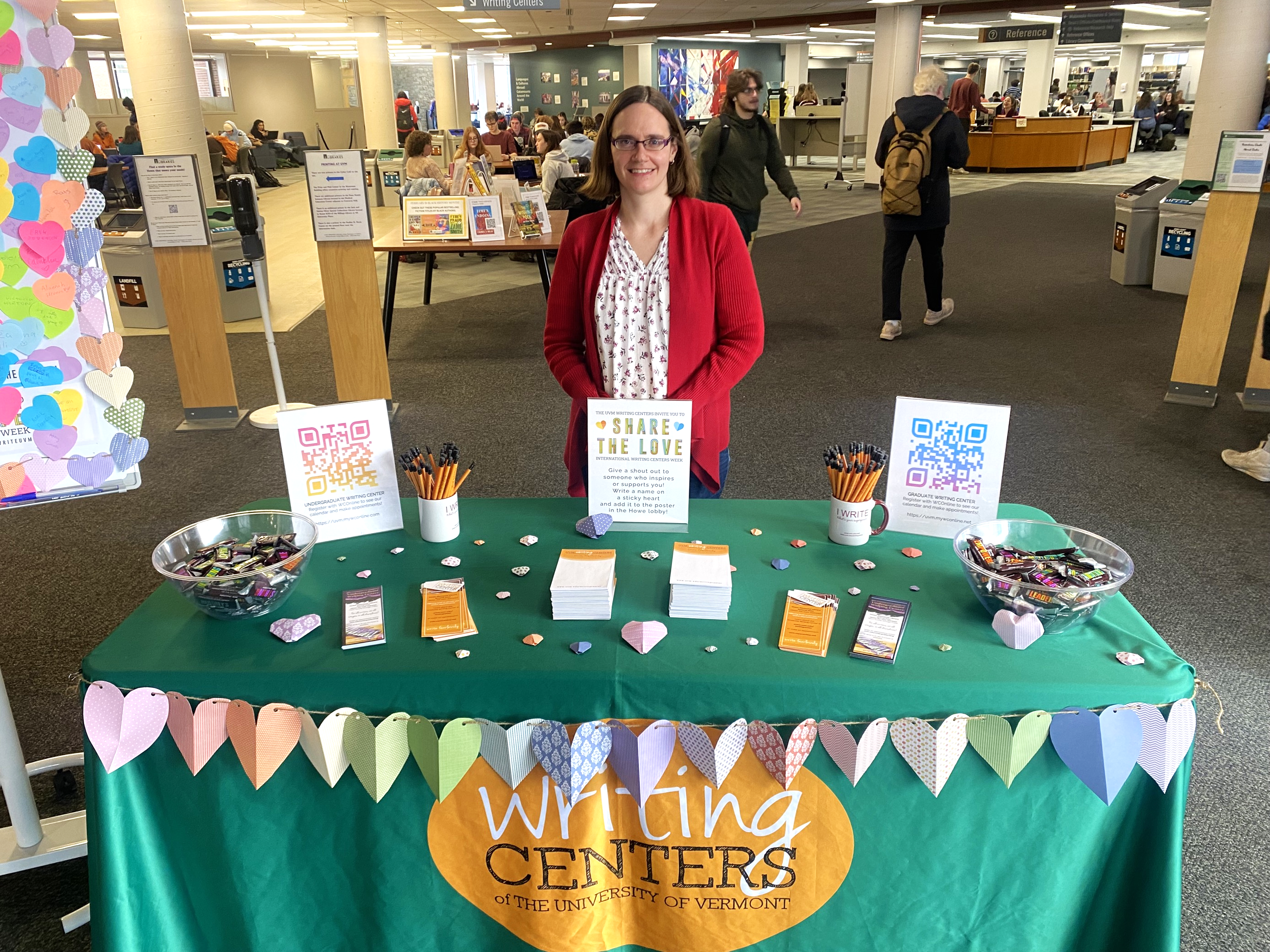 April Christenson stands behind a table draped with the writing centers' table cloth. The table displays two bowls of chocolates, origami hearts, and QR codes. A banner of paper hearts is strung across the front of the tablecloth.