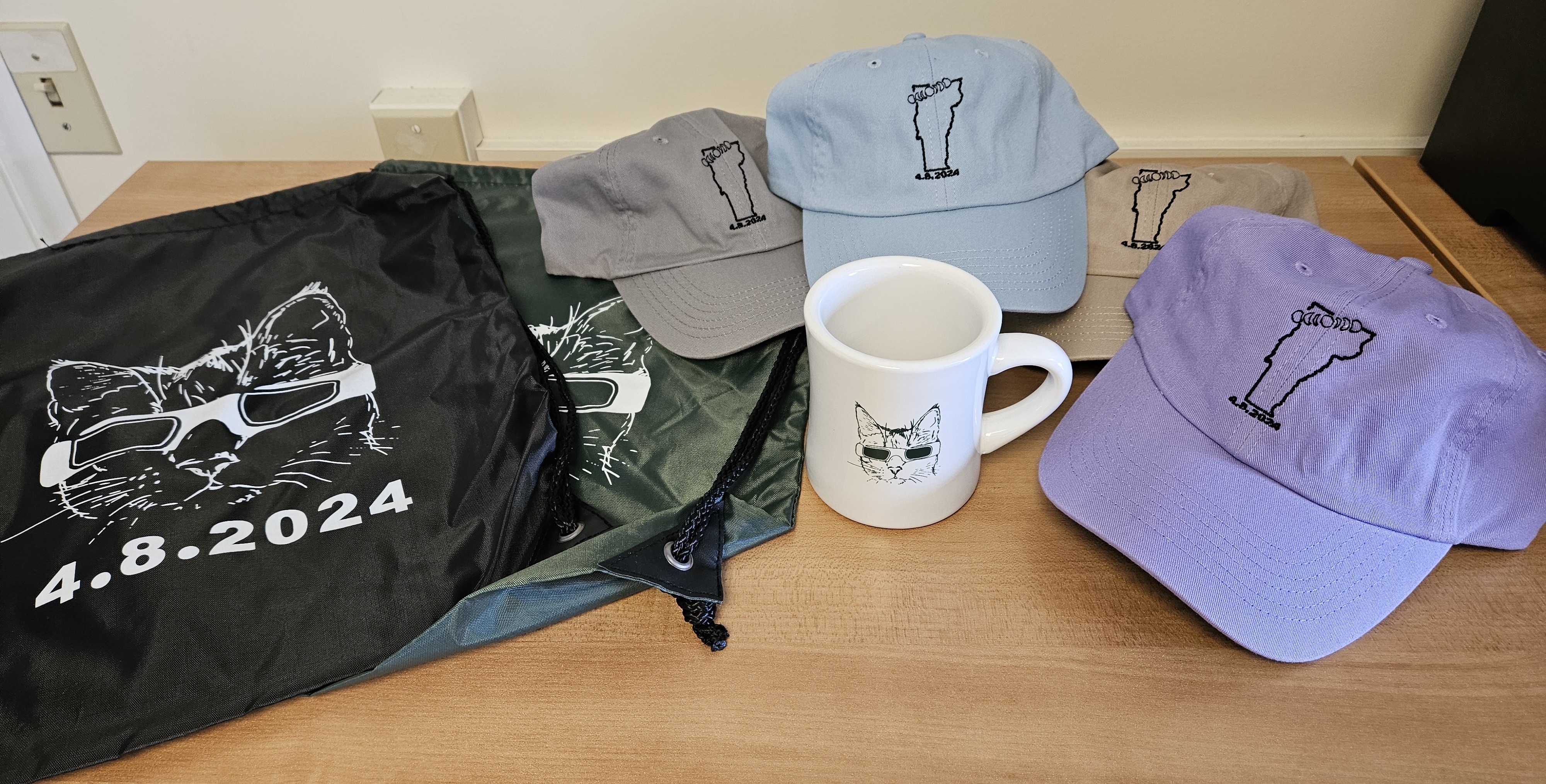 eclipse swag showing drawstring bags with the eclipse cat, baseball hats with the eclipse path embroidered on them, and a diner mug with the eclipse cat.